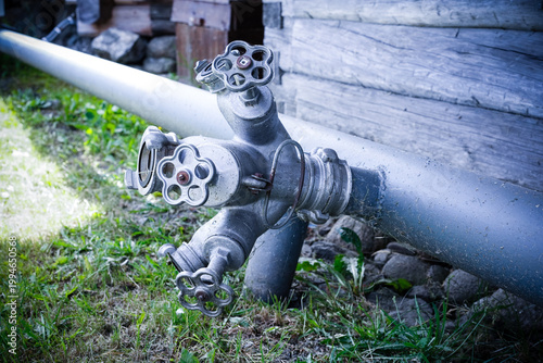 Industrial metal pipe assembly with multiple control valves and handles outdoors on grass next to a wooden building wall.