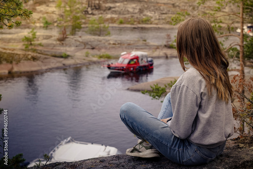 A young woman sits on a high rocky ledge, her back to the camera, overlooking a calm bay where a red motorboat is cruising through a rugged, northern landscape. Sortavala, Karelia, skerries, Ladoga