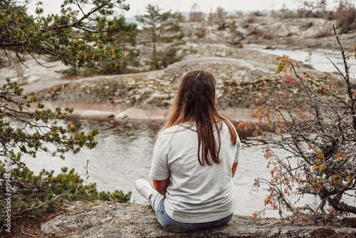 A woman sits on a rocky cliff, looking out over a calm river and a wilderness landscape of stone and scattered evergreens of Karelia