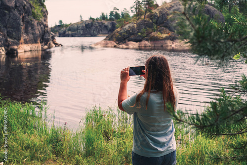 A woman stands at the grassy edge of a calm bay, using her smartphone to photograph a rugged northern landscape of granite cliffs and evergreen trees.