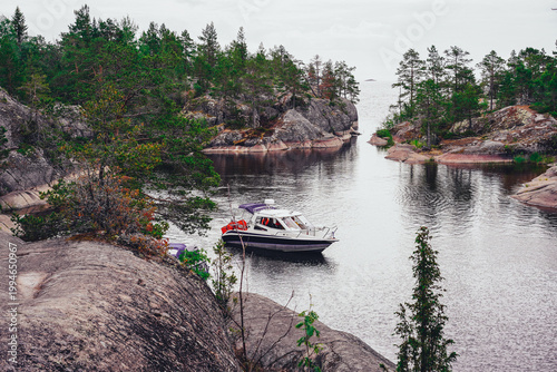 A scenic view of a motorboat moving across a calm lake, leaving a gentle wake, surrounded by rugged rocky shores and a dense evergreen forest under a soft, overcast sky. Karelia
