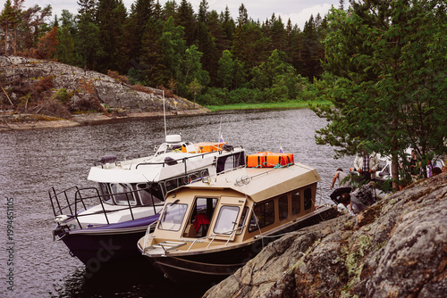 Two passenger motorboats are moored against a rocky shore in a rugged northern landscape Karelia, with dense evergreen forests and rocky hills in the background.