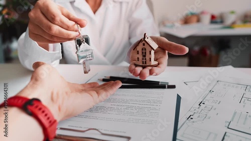 Person giving house keys over paperwork with small home model in background. Symbol of apartment sale, rental agreement, successful closing deal and secure housing investment process.