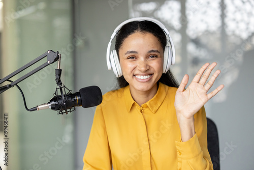 Smiling woman wearing headphones and waving while sitting at a desk with a professional microphone, recording audio for a podcast or streaming an online show