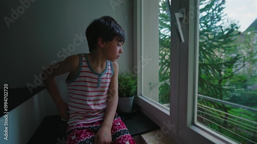 Young child standing by open window looking outside at trees, thoughtful expression and quiet moment of curiosity, natural light entering room creating calm reflective indoor atmosphere