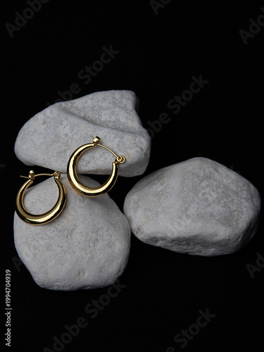 Elegant gold hoop earrings resting on smooth white stones against a black background — minimalist jewelry shot