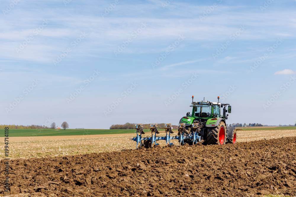 Fototapeta premium Modern tractor plowing agricultural field in spring, soil cultivation with plow, farming machinery working on farmland under blue sky