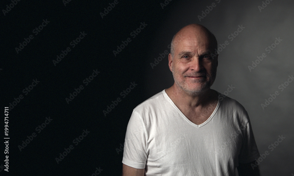 Obraz premium Smiling mature adult man in white t-shirt on dark background looking at camera