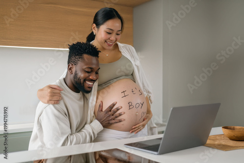 Happy multicultural couple expecting a baby sharing a joyful video call with family. Pregnant asian woman and african partner smiling at laptop showing grandparents. Warm home, love, connection