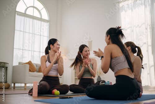 Group of young Asian women seated in a circle on yoga mats in modern living room, laughing and clapping together after a wellness class.