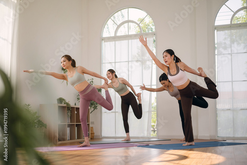 Group of young healthy Asian women practicing yoga class in home studio. Active women performing standing balance and stretch on yoga mats. Concept of healthy lifestyle.