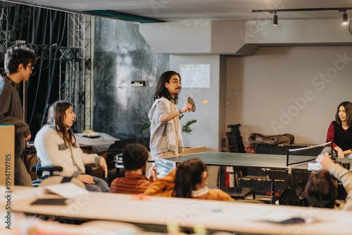 Students play a friendly game of table tennis around a ping-pong table. A diverse group watches and socializes in a relaxed indoor recreation setting.