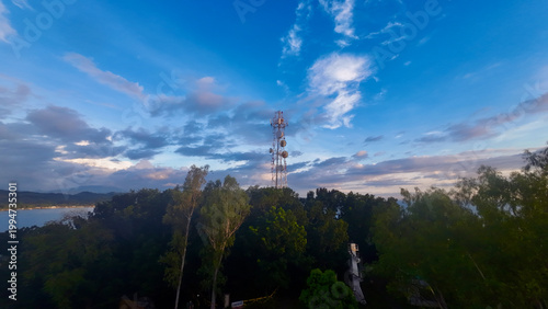 Telecommunication tower rising above tropical forest at sunset. Modern antenna structure against dramatic sky with clouds and warm evening light.