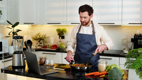 Middle-aged man smiles while seasoning vegetables in pan on electric stove for healthy diet meal. Young guy watches laptop recipe video while cooking colorful food in home kitchen indoors for fitness.