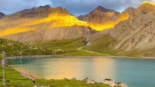 Numerous mountains with the sun setting, casting warm light on the peak and creating a reflection on the water of Tian Tang Lake in Xinjiang Province, China. Lush green grasslands form in foreground.