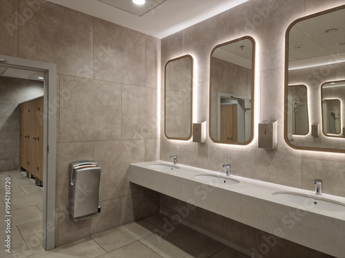 Modern public restroom with backlit mirrors, stone tiles, and white integrated vanity sinks