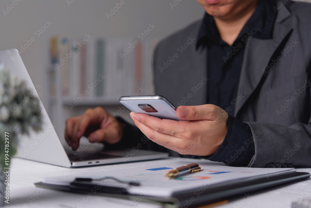 Fototapeta premium Businessman hand holding smartphone typing laptop computer desk blurred financial chart corporate office multitasking digital communication cross device workflow synchronization efficiency.