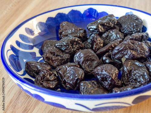 Dried prunes in a decorative ceramic bowl on a wooden table, close-up view. Healthy natural snack rich in fiber and antioxidants, rustic food background with selective focus