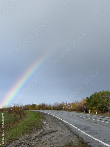A beautiful rainbow arcs over a tranquil road, framed by autumn foliage and a calm sky.