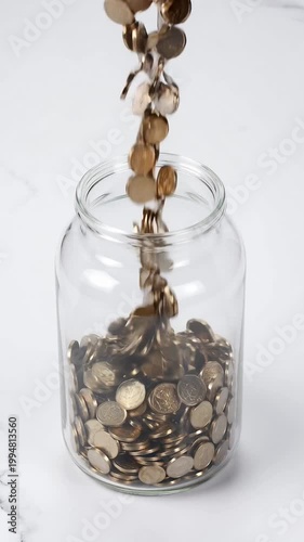 Gold Coins Falling into Clear Glass Jar on White Marble Surface Under Bright Lighting Saving Money Concept