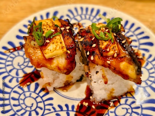 High angle view of two unagi nigiri sushi topped with green onion and sesame seeds, served on patterned ceramic plate