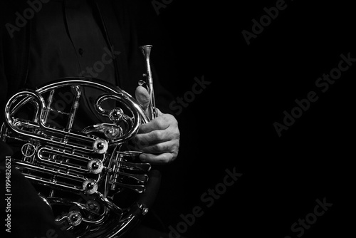  A close-up of a French horn in the hands of a musician in black and white against a black background