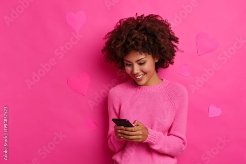 Woman in Pink Sweater Smiles While Using Smartphone Against Pink Wall With He...