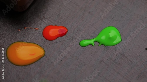 Close-up of vibrant paint droplets in orange, green, and yellow on a textured surface with a brush applying green paint.
