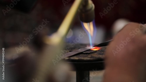 A close-up of a jeweler using a blowtorch to heat a metal piece, showcasing the craftsmanship of metalworking with glowing embers and focused flame.