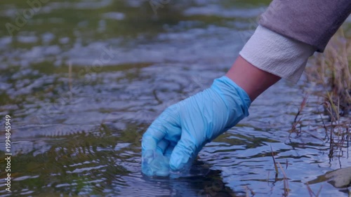 Female scientist wearing blue protective gloves collecting a water sample from a stream in a test tube to measure pollution levels and water quality, an important part of environmental analysis
