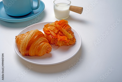Coissant on a white plate, with a coffee cup and a pitcher of fresh milk in the background.