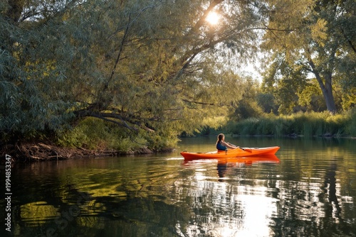 Woman Paddles Kayak on Calm River Surrounded by Trees in Late Afternoon Sunlight
