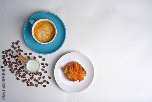 Croissant on a white plate, with a coffee cup and a pitcher of fresh milk in the background.