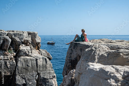 A mother and daughter are sitting on a rocky cliff and looking at the sea. 