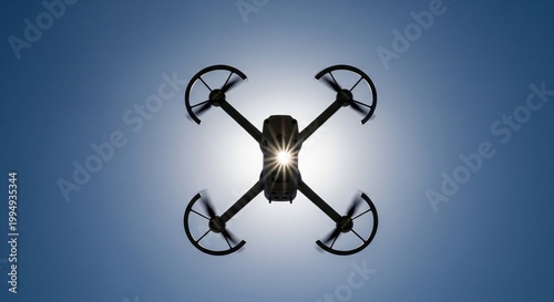 Dark quadcopter drone flying high in clear blue sky against bright sun viewed from below
