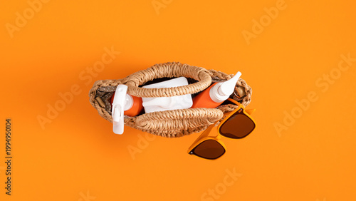 Top View of Summer Essentials in Woven Straw Bag on Bright Orange Background. Flat Lay with Sunscreen Spray Bottles and Sunglasses. Concept of Beach Vacation, Skin Care, SPF and Travel Planning.