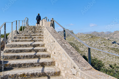 Wide shot of the concrete and stone staircase with metal handrails at a scenic mountain viewpoint overlooking the rugged landscape of the Balearic Islands.