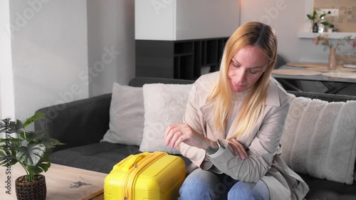 Worried and pensive woman sit at home, nervously check smartwatch with suitcase and keys on table. Concept of emotional life decision, relationship uncertainty, and major personal transition.