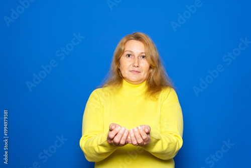 A blonde Caucasian woman in her forties offers something with a friendly gesture, isolated against a blue studio background