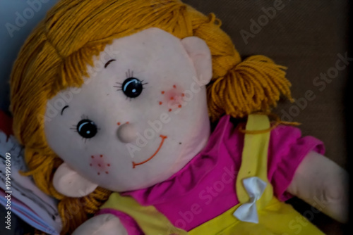 Szmaciana lalka z uśmiechniętą buzią i włóczkowymi włosami. Ma czarne, guzikowate oczy i pomarańczowe, wyszywane usta. Włosy z żółtej włóczki, upięte w prosty warkoczyk z boku. 