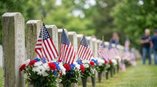 American flags and floral arrangements placed at gravestones in a cemetery during Memorial Day observance, with visitors in the background honoring fallen soldiers