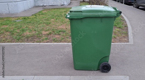 Large green plastic garbage bin stands on a paved sidewalk in a residential area. Clean waste container. For themes related to urban ecology, recycling, and city waste management systems