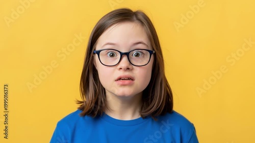 Portrait of a young girl with Down syndrome. Happy child with glasses looking surprised on yellow background. Inclusion and diversity concept