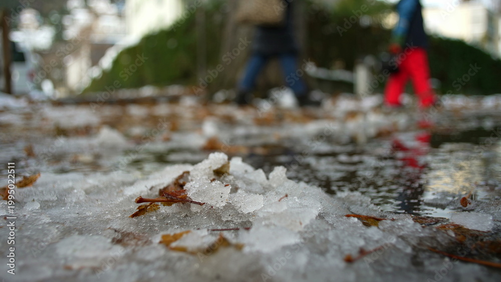 Naklejka premium Melting snow on city sidewalk with fallen leaves and reflections, subtle sign of seasonal change as people walk in background, early winter thaw and urban transition