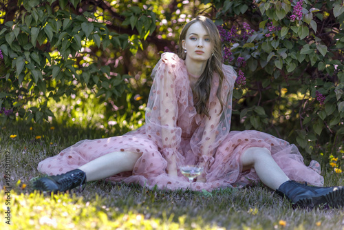 A portrait of a young woman with long hair in a pink dress among blooming lilacs. She sits with a glass of wine, relaxing on a picnic. Spring blossoms.