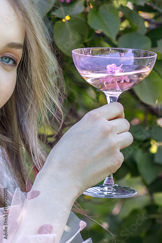 A portrait of a young woman with long hair in a pink dress among blooming lilacs. She sits with a glass of wine, relaxing on a picnic. Spring blossoms.