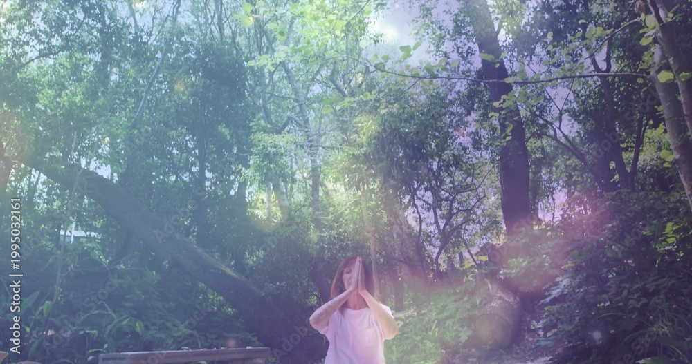 Fototapeta premium Standing woman wearing white T-shirt holding hands in forest glade with bench fallen trunk sunbeams