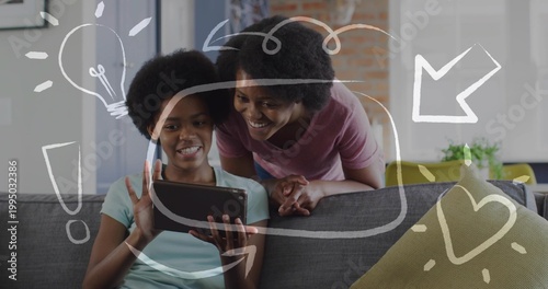 Smiling mother and daughter leaning over gray sofa at home, holding tablet with white doodles