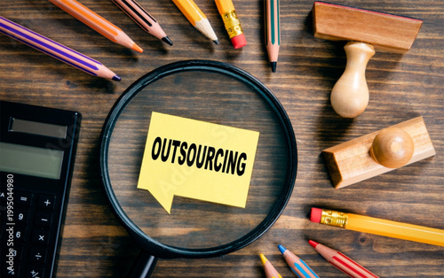 Outsourcing concept with magnifying glass on wooden desk surrounded by office supplies