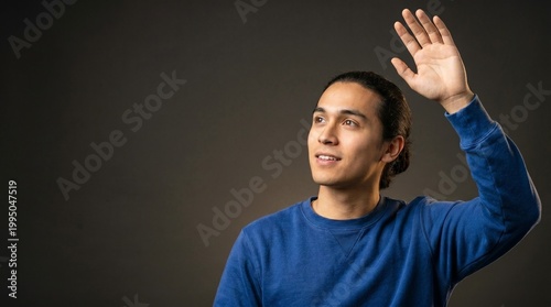 Young man waving hand in greeting gesture. Guy smiling and showing waving motion with arm. Friendly person communicating with hello sign. Casual lifestyle portrait against dark background.